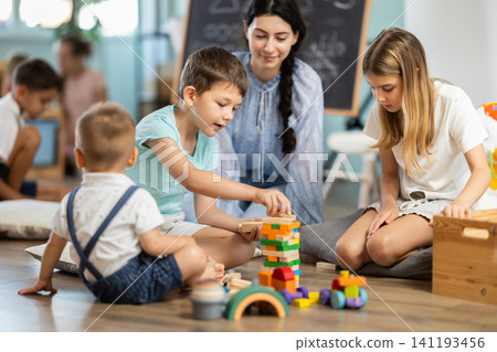 Children playing in living room of kindergarten, babysitter support and participate in game. Children playing in living room of kindergarten, babysitter support and participate in game. 141193456