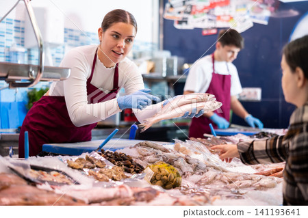Female seller in apron standing near counter offering fresh fish striped red mullet to buyer 141193641