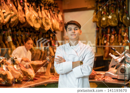 Portrait of experienced butcher on background of rack with hanging various jamon Portrait of experienced butcher on background of rack with hanging various jamon 141193670