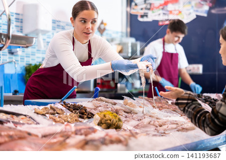 Portrait of saleswoman in apron offering female customer fresh fish at shop Portrait of saleswoman in apron offering female customer fresh fish at shop 141193687