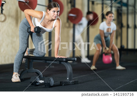 Athletic young woman pumping her biceps with a dumbbell while leaning on bench Athletic young woman pumping her biceps with a dumbbell while leaning on bench 141193716