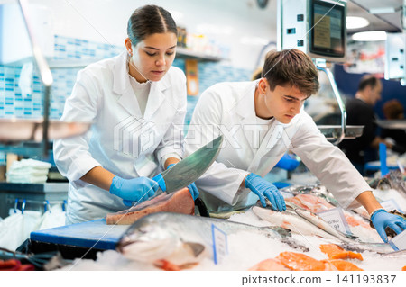 Female seller with knife cut fish in supermarket near counter Female seller with knife cut fish in supermarket near counter 141193837