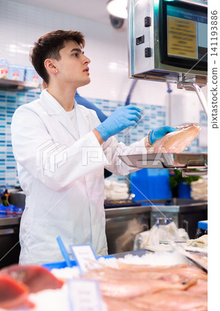 Young seller in fish department of supermarket weighs atlantic cod fish on scale Young seller in fish department of supermarket weighs atlantic cod fish on scale 141193886