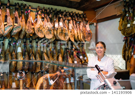 Young female butcher shopkeeper with crossed arms near hanging legs of Iberian ham Young female butcher shopkeeper with crossed arms near hanging legs of Iberian ham 141193906