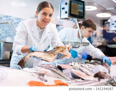 Positive saleswoman holding cod fish in fish store 141194087