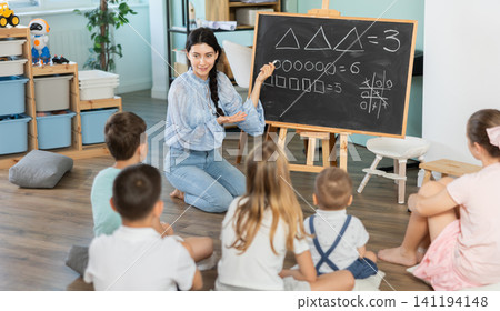 teacher in a kindergarten stands at the blackboard in front of children 141194148
