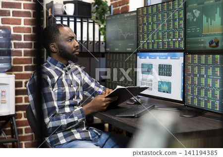 Man holding clipboard inside trading office as market screens, documents and charts support financial analysis in a trading office. Man holding clipboard inside trading office as market screens, documents and charts support financial analysis in a trading office. 141194385