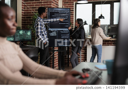 Backend network developer explains application code to a coworker as the team builds security layers for stable software functionality. Backend network developer explains application code to a coworker as the team builds security layers for stable software functionality. 141194399