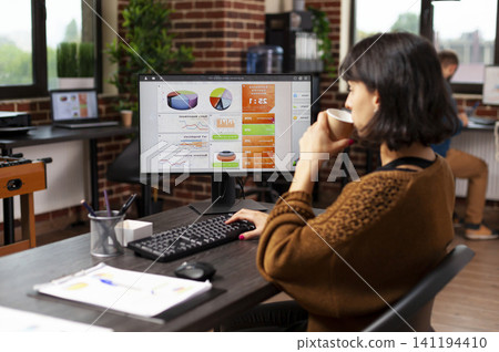 Businesswoman holds coffee while studying financial graphs at her desk, working through marketing strategy and company project analysis. Businesswoman holds coffee while studying financial graphs at her desk, working through marketing strategy and company project analysis. 141194410