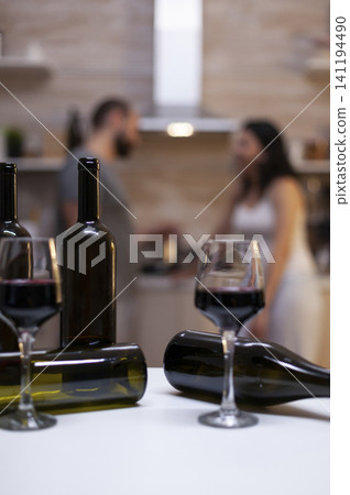 Empty bottles and wine glasses stand on a kitchen counter while an intoxicated couple sits behind them, showing alcohol abuse and unhealthy home drinking. Empty bottles and wine glasses stand on a kitchen counter while an intoxicated couple sits behind them, showing alcohol abuse and unhealthy home drinking. 141194490