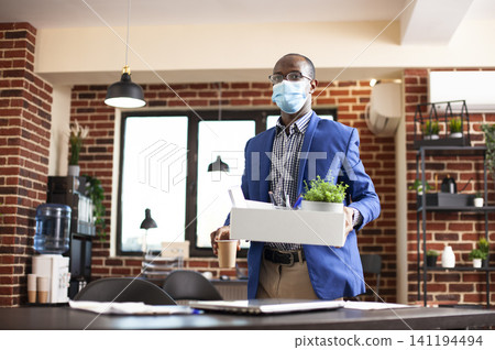 Masked employee carries a white box and coffee cup toward an office desk, suggesting work belongings, relocation and professional transition. Masked employee carries a white box and coffee cup toward an office desk, suggesting work belongings, relocation and professional transition. 141194494