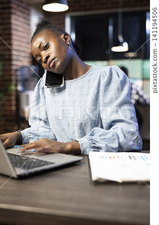 Black entrepreneur talks on a smartphone while entering financial information into a laptop, multitasking during night stock market analysis. 141194506