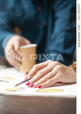 Woman points with a pen at financial documents near coffee on a table, emphasizing paperwork review, selective focus and company research in an office task. 141194507