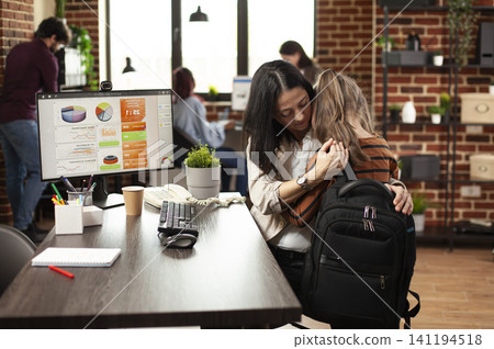 Working mother hugs her sleepy daughter at a startup desk with monitor, school bag and notebook, blending childcare with office responsibilities. Working mother hugs her sleepy daughter at a startup desk with monitor, school bag and notebook, blending childcare with office responsibilities. 141194518