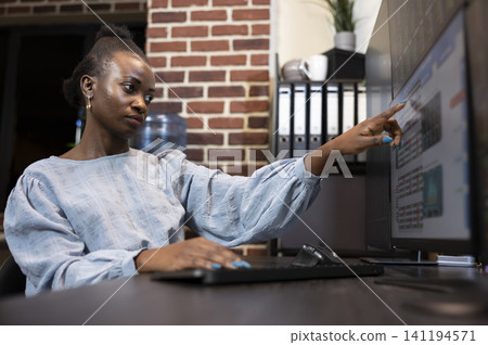 Black female finance analyst points at computer screens while monitoring forex data, hedge fund information and stock market movements, with chart analysis and trading workspace context. Black female finance analyst points at computer screens while monitoring forex data, hedge fund information and stock market movements, with chart analysis and trading workspace context. 141194571