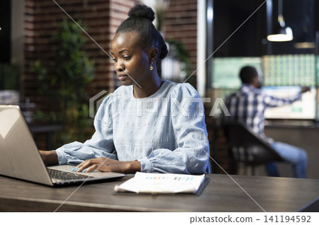 Focused Black woman reviews reports on a laptop during a late night office shift while a colleague works in the background near financial charts. 141194592
