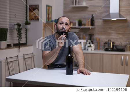 Pensive man drinking glass of wine while sitting alone and depressed, person with alcohol addiction having bottle of alcoholic beverage, liquor, booze. Pensive man drinking glass of wine while sitting alone and depressed, person with alcohol addiction having bottle of alcoholic beverage, liquor, booze. 141194596