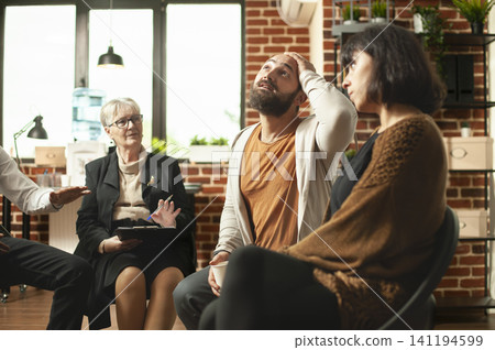 Bearded man holds his head while speaking in a group therapy circle as a senior therapist offers support during counseling for emotional struggles. Bearded man holds his head while speaking in a group therapy circle as a senior therapist offers support during counseling for emotional struggles. 141194599