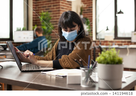 Focused businesswoman reads company documents and research data on a laptop at an office desk while wearing a protective face mask. Focused businesswoman reads company documents and research data on a laptop at an office desk while wearing a protective face mask. 141194618