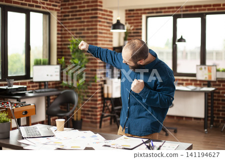 Cheerful businessman raises his fists in a startup office after project success, expressing victory, motivation and confidence in business planning. Cheerful businessman raises his fists in a startup office after project success, expressing victory, motivation and confidence in business planning. 141194627