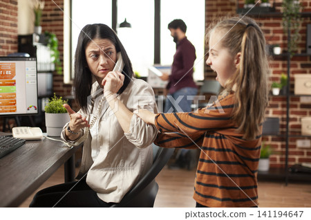 Mother at an office desk tries to speak with a client by phone while her daughter pulls her arm and financial charts remain open on the computer. Mother at an office desk tries to speak with a client by phone while her daughter pulls her arm and financial charts remain open on the computer. 141194647