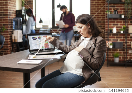 Pregnant businesswoman reviewing research reports showing pregnant businesswoman sitting and reviewing research data at office desk with laptop nearby. Pregnant businesswoman reviewing research reports showing pregnant businesswoman sitting and reviewing research data at office desk with laptop nearby. 141194655