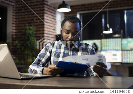 Focused Black businessman sits at a desk holding a clipboard while monitoring forex trading trends and stock market data on a nearby laptop. Focused Black businessman sits at a desk holding a clipboard while monitoring forex trading trends and stock market data on a nearby laptop. 141194668