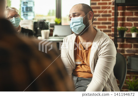 Sad man wearing a face mask speaks about anxiety and distress during an addiction counseling session with a support group in a brick wall room. Sad man wearing a face mask speaks about anxiety and distress during an addiction counseling session with a support group in a brick wall room. 141194672