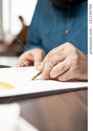Hand of employee holding pen over financial paperwork on table, with closeup of caucasian person writing and reviewing company research data on clipboard in startup office. Hand of employee holding pen over financial paperwork on table, with closeup of caucasian person writing and reviewing company research data on clipboard in startup office. 141194708