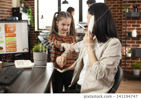 Businesswoman in startup office struggles to focus on client call, distracted by playful daughter disrupting workplace, Little child pulling arm of her working mom talking on phone in brick wall room. Businesswoman in startup office struggles to focus on client call, distracted by playful daughter disrupting workplace, Little child pulling arm of her working mom talking on phone in brick wall room. 141194747