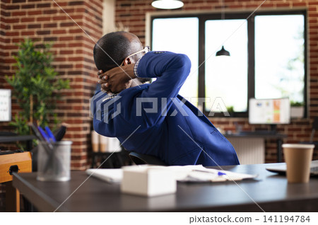 Manager relaxing at his desk, leaning back in chair and taking moment to unwind in startup office, Black businessman with hands behind his head, pausing to relax in modern brick wall workspace. Manager relaxing at his desk, leaning back in chair and taking moment to unwind in startup office, Black businessman with hands behind his head, pausing to relax in modern brick wall workspace. 141194784