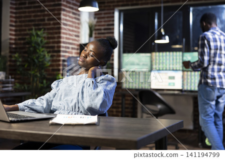 African american woman experiencing work fatigue massages her neck, taking a break from analyzing data on her laptop, the nighttime setting emphasizes the intensity of late shifts. African american woman experiencing work fatigue massages her neck, taking a break from analyzing data on her laptop, the nighttime setting emphasizes the intensity of late shifts. 141194799
