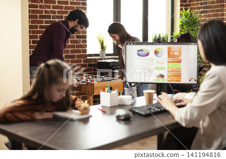 Businesswoman reviews market analysis on desktop pc, sharing desk with daughter as male and female coworkers play foosball, with mother and child seated at table as her colleagues relieve stress in. Businesswoman reviews market analysis on desktop pc, sharing desk with daughter as male and female coworkers play foosball, with mother and child seated at table as her colleagues relieve stress in. 141194816