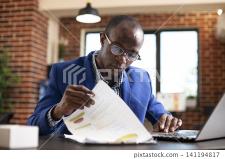 Black man seated at office desk, deeply focused on analyzing financial documents in brick wall room, with african american male manager examining graphs on clipboard with laptop nearby. Black man seated at office desk, deeply focused on analyzing financial documents in brick wall room, with african american male manager examining graphs on clipboard with laptop nearby. 141194817