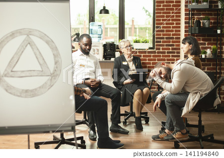 Group of alcoholics gather in aa meeting room with symbol displayed on white board. Retired therapist conducting supportive conversation, discussing mental health and addiction in brick wall office. Group of alcoholics gather in aa meeting room with symbol displayed on white board. Retired therapist conducting supportive conversation, discussing mental health and addiction in brick wall office. 141194840