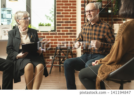 Older man shares personal struggles during addiction recovery as a senior female therapist listens carefully and takes notes in a supportive group therapy session. Older man shares personal struggles during addiction recovery as a senior female therapist listens carefully and takes notes in a supportive group therapy session. 141194847