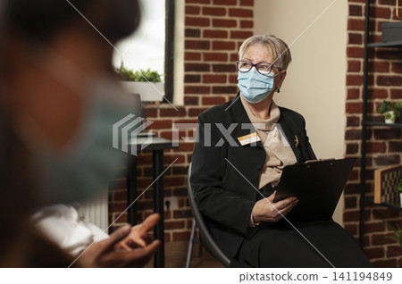 Woman therapist listens to people in an addiction support meeting while wearing a protective mask and offering counseling guidance in a group therapy room. Woman therapist listens to people in an addiction support meeting while wearing a protective mask and offering counseling guidance in a group therapy room. 141194849