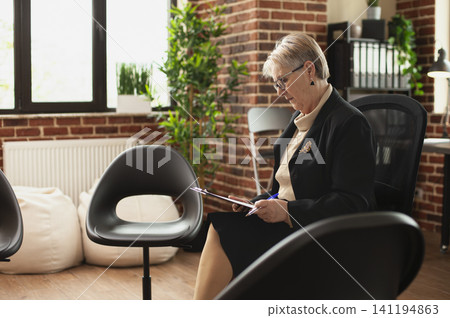 Senior psychiatrist in glasses reviews clipboard notes among arranged chairs in a brick wall therapy room prepared for mental health counseling. Senior psychiatrist in glasses reviews clipboard notes among arranged chairs in a brick wall therapy room prepared for mental health counseling. 141194863