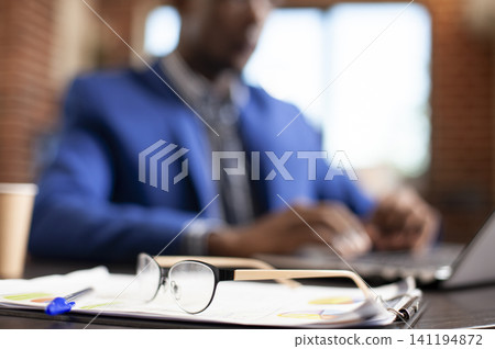 Spectacles and clipboard sit on a desk while African American employee uses laptop in background, eyeglasses, paper documents and pen positioned on table for businessman to use in startup office. Spectacles and clipboard sit on a desk while African American employee uses laptop in background, eyeglasses, paper documents and pen positioned on table for businessman to use in startup office. 141194872