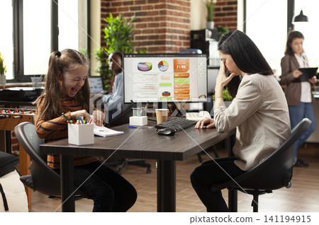 Noisy daughter interrupts mother during office work at a startup office table, showing a working mother distracted by her child while trying to use a computer. Noisy daughter interrupts mother during office work at a startup office table, showing a working mother distracted by her child while trying to use a computer. 141194915