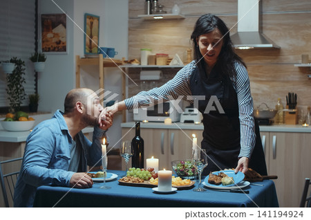 Thoughtful wife serves her husband during a romantic kitchen dinner, sharing healthy food and conversation at the anniversary table, inside a realistic lifestyle setting. 141194924
