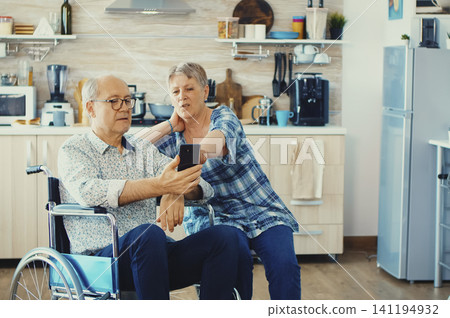 Senior woman and disabled husband in a wheelchair browse the internet on a smartphone in the kitchen using modern communication technology. Senior woman and disabled husband in a wheelchair browse the internet on a smartphone in the kitchen using modern communication technology. 141194932