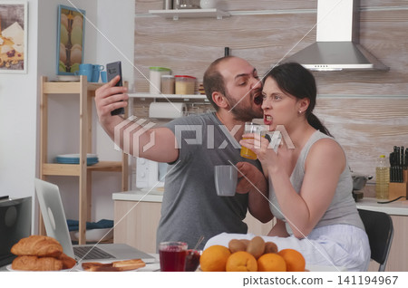 Happy young couple take selfies during breakfast in the kitchen, making funny faces and sharing a cheerful married lifestyle moment. 141194967