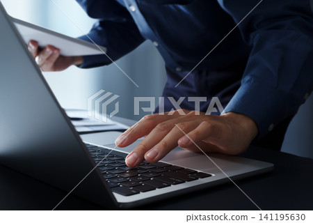 一位商務人士坐在辦公桌前使用電腦，同時看著平板電腦。鏡頭特寫他的雙手和鍵盤。商務科技 141195630