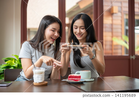 Asian women friends looking at smartphone together while enjoying coffee and dessert in a cozy cafe. Asian women friends looking at smartphone together while enjoying coffee and dessert in a cozy cafe. 141195689