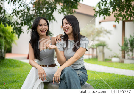 Two attractive Asian women sitting on a rock in the garden, talking, gesturing and laughing together happily. Two attractive Asian women sitting on a rock in the garden, talking, gesturing and laughing together happily. 141195727