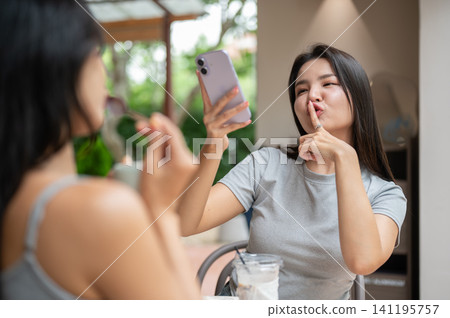 Funny Asian woman holding smartphone up high while making a hushing finger gesture at a cafe. Funny Asian woman holding smartphone up high while making a hushing finger gesture at a cafe. 141195757