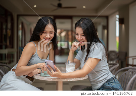 Asian female friends smiling while browsing smartphone together over coffee at outdoor cafe terrace. Asian female friends smiling while browsing smartphone together over coffee at outdoor cafe terrace. 141195762