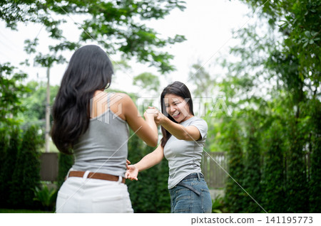 Two happy Asian women holding hands and spinning around on a green lawn. 141195773
