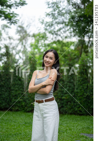 Asian woman standing confidently in a green garden with folded arms and a cheerful smile outdoors. Asian woman standing confidently in a green garden with folded arms and a cheerful smile outdoors. 141195783